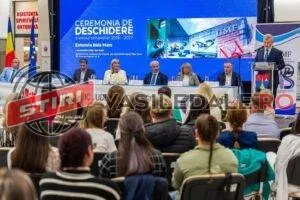 Un grup de persoane stă așezat în scaune, privind spre un podium unde mai multe persoane sunt poziționate în fața unui ecran mare albastru.