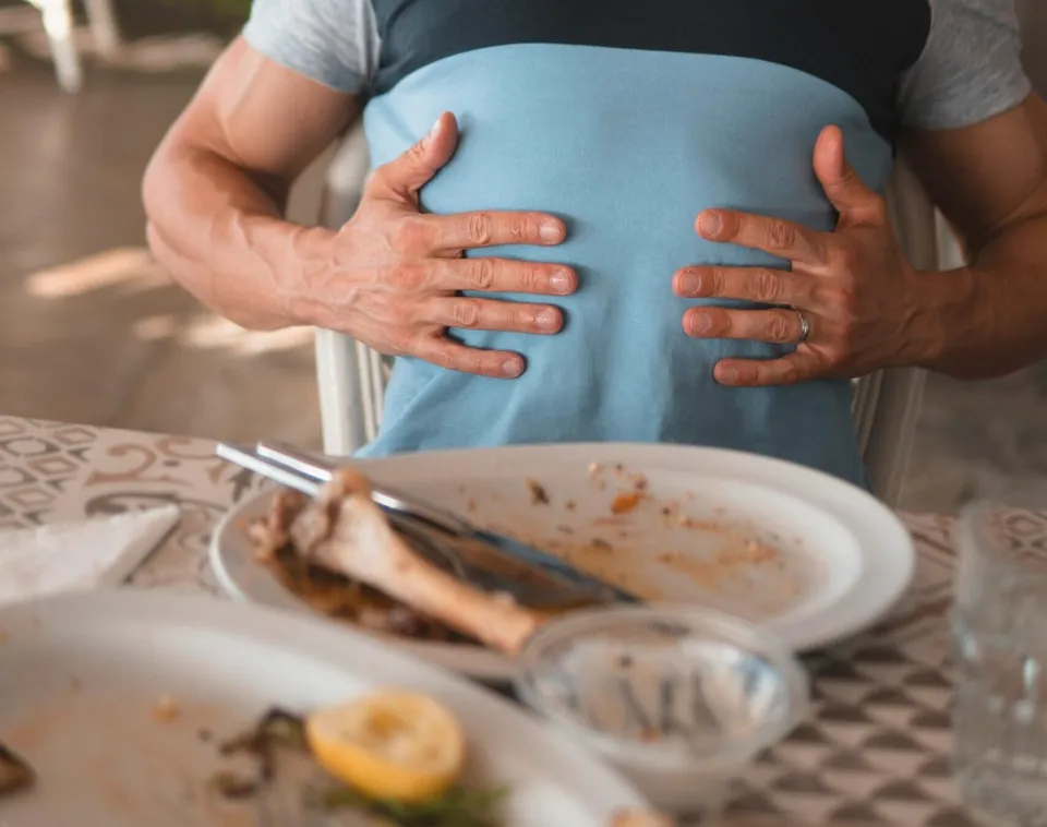 Un bărbat cu tricou albastru își ține mâinile pe abdomen, stând la o masă cu farfurii cu resturi de mâncare și un fel de lămâie.