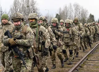 Un grup de soldați în uniforme de camuflaj și echipament militar merge pe lângă o cale ferată într-un teren deschis.