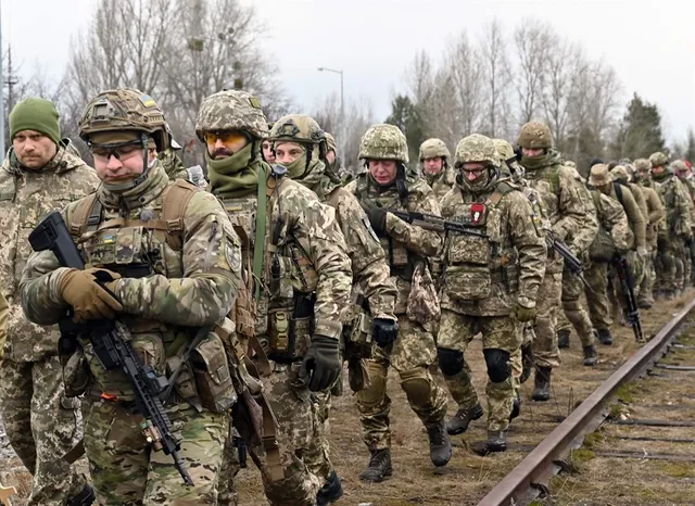 Un grup de soldați în uniforme de camuflaj și echipament militar merge pe lângă o cale ferată într-un teren deschis.
