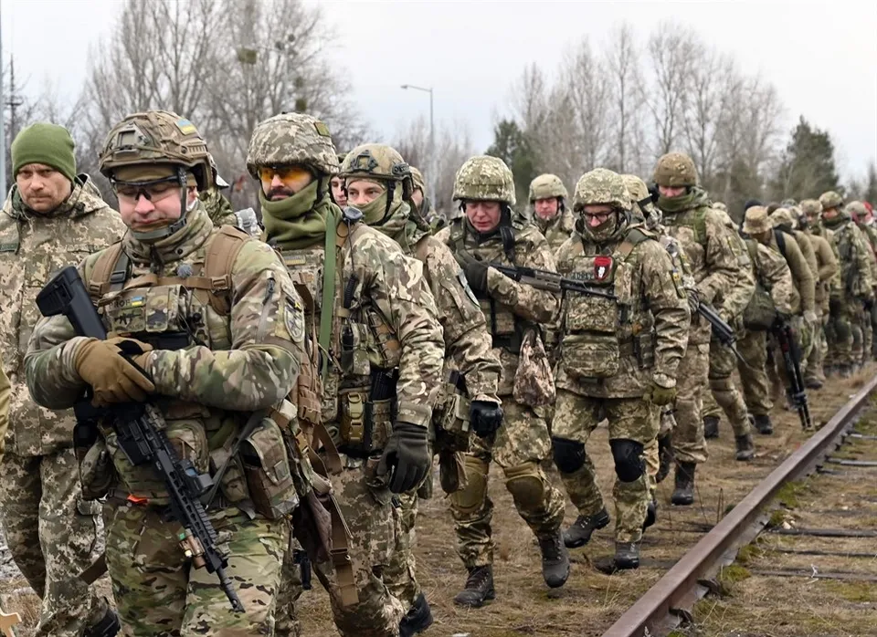 Un grup de soldați în uniforme de camuflaj și echipament militar merge pe lângă o cale ferată într-un teren deschis.