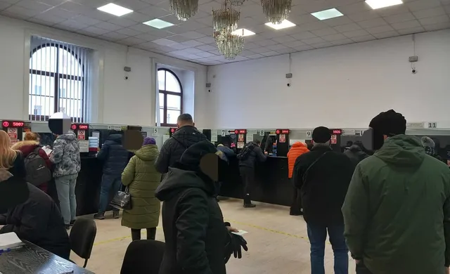 Oamenii stau în rând în fața unor birouri negre într-o încăpere mare cu pereți albi și plafon cu lumini încorporate.