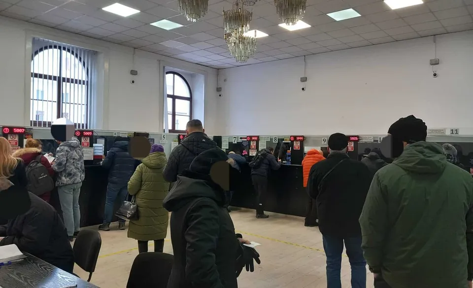 Oamenii stau în rând în fața unor birouri negre într-o încăpere mare cu pereți albi și plafon cu lumini încorporate.