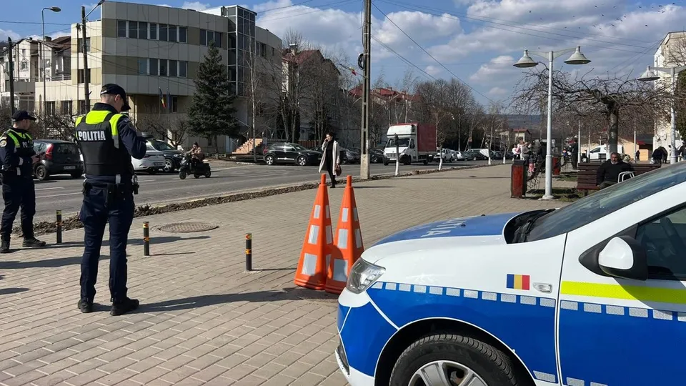 Un polițist stă pe un trotuar lângă un vehicul de serviciu, având conuri de trafic portocalii și clădiri în fundal.