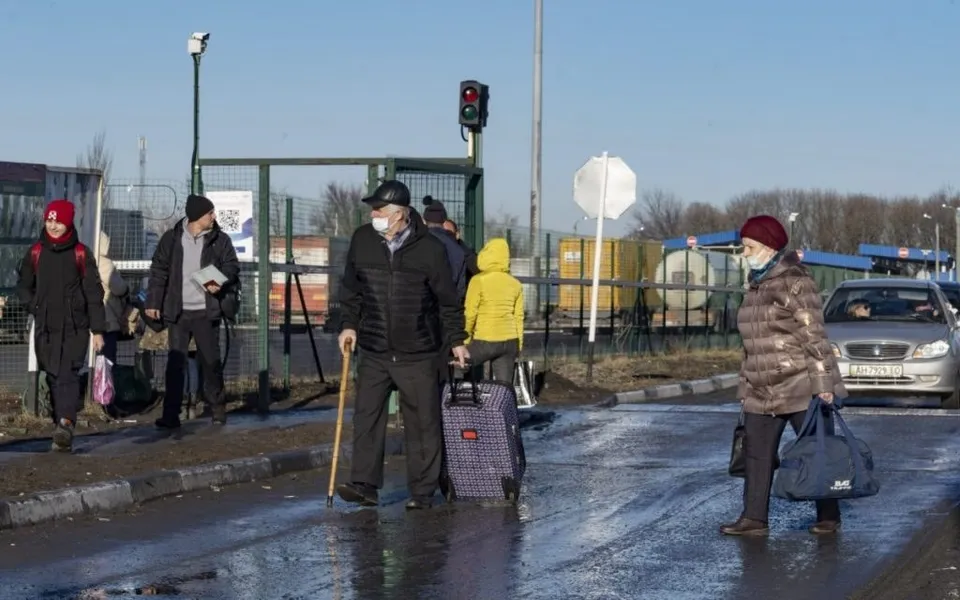 Oameni cu bagaje merg pe un drum asfaltat lângă un gard metalic sub un cer senin.