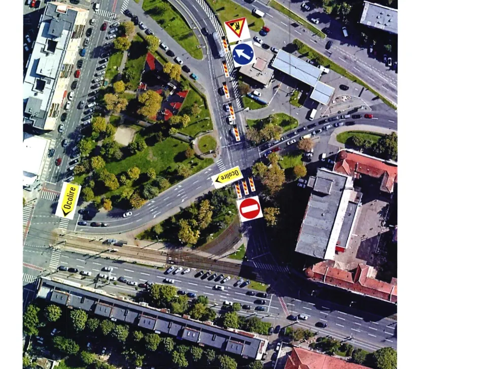 O vedere aeriană a unei intersecții urbane cu mașini parcate, drumuri asfaltate, spații verzi și câteva indicatoare rutiere de circulație.
