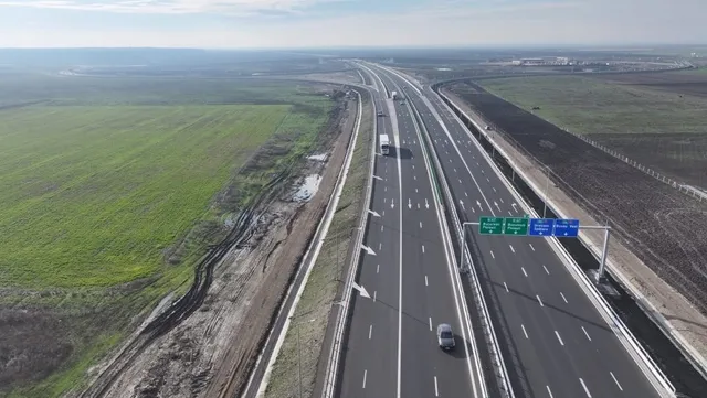 O vedere aeriană a unei autostrăzi cu mai multe benzi de circulație care traversează un peisaj deschis cu câmpuri verzi și zone de teren pregătit.