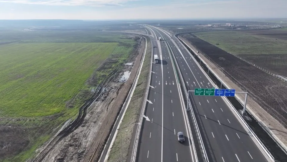 O vedere aeriană a unei autostrăzi cu mai multe benzi de circulație care traversează un peisaj deschis cu câmpuri verzi și zone de teren pregătit.