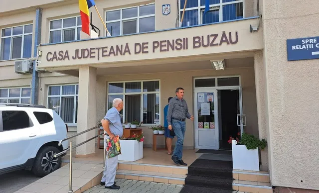 Un bărbat cu părul alb stă lângă o rampă de acces, în timp ce un alt bărbat coboară scările unei clădiri cu inscripția Casa Județeană de Pensii Buzău.
