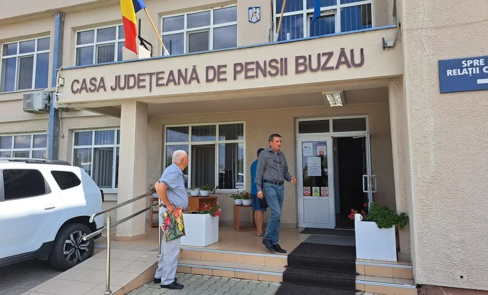 Un bărbat cu părul alb stă lângă o rampă de acces, în timp ce un alt bărbat coboară scările unei clădiri cu inscripția Casa Județeană de Pensii Buzău.