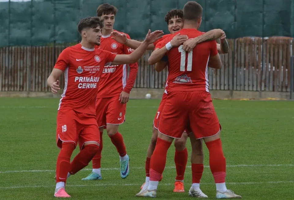 Cinci jucători de fotbal îmbrăcați în echipamente roșii sărbătoresc pe un teren de gazon verde.