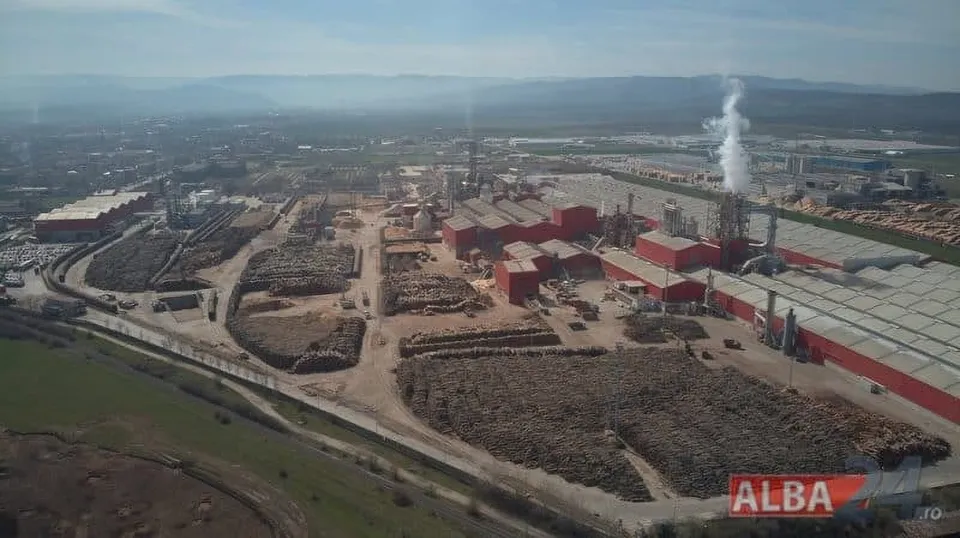 O vedere aeriană a unui complex industrial cu clădiri roșii, depozite de materiale și coloane de fum alb ce se ridică spre cer.