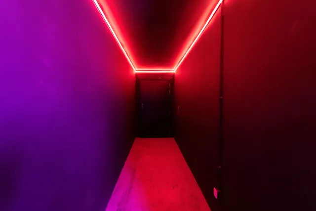 Un coridor îngust cu pereți și podea iluminate de benzi de lumină roșie și violet care duc spre o ușă întunecată în fundal.