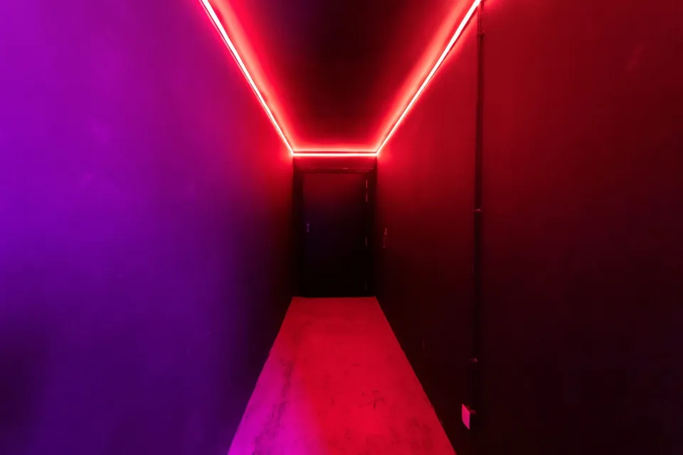 Un coridor îngust cu pereți și podea iluminate de benzi de lumină roșie și violet care duc spre o ușă întunecată în fundal.