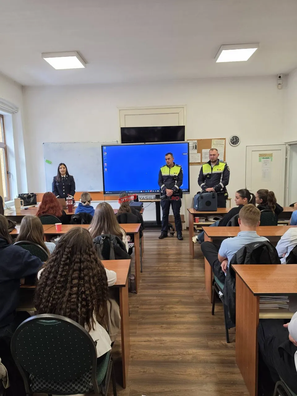 În fața unui ecran mare, doi bărbați în uniforme de polițist stau în picioare într-o sală de clasă în fața unor elevi așezați la bănci.