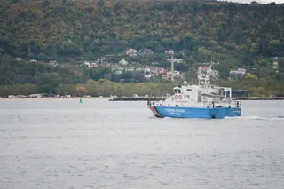 O barcă albastră și albă cu inscripții pe lateral navighează pe o suprafață de apă, având un deal împădurit cu case în fundal.