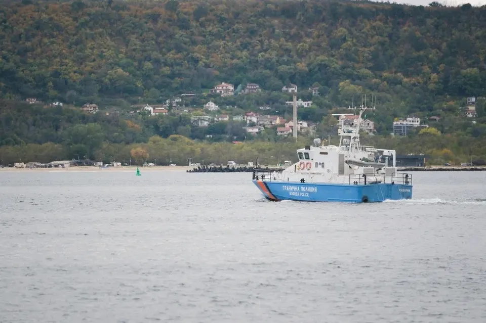 O barcă albastră și albă cu inscripții pe lateral navighează pe o suprafață de apă, având un deal împădurit cu case în fundal.