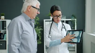 O femeie cu halat alb și stetoscop arată unui bărbat cu ochelari imagini medicale afișate pe o tabletă digitală.