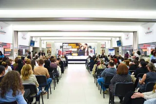 Mulți oameni stau pe scaune dispuse în rânduri, orientate spre un panou informativ situat în partea de fund a unui hol spațios.