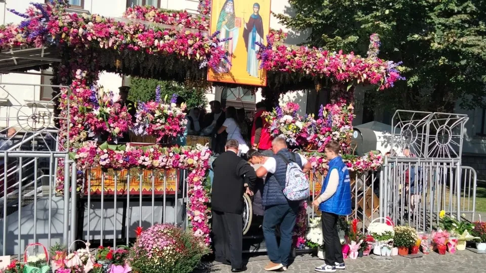 Un grup de persoane stă lângă un vehicul decorat abundent cu flori roz și mov, lângă un gard metalic și plante verzi.
