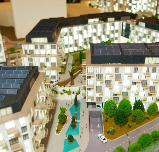 O machetă arhitecturală prezintă clădiri de apartamente albe cu ferestre și panouri solare pe acoperiș, dispuse pe o suprafață cu zone verzi și drumuri.