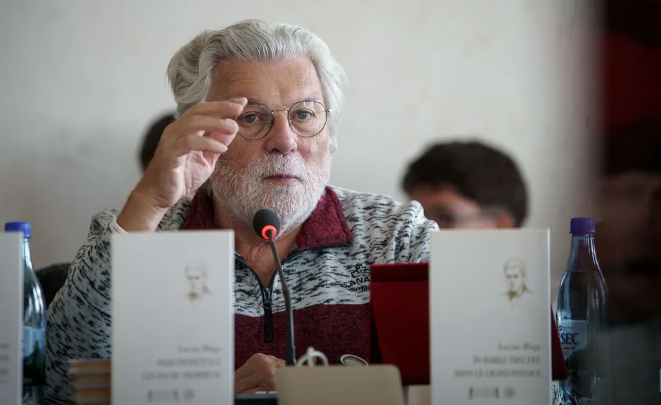 Un bărbat cu părul alb și ochelari își ajustează lentila, stând la o masă cu cărți și microfon în față.