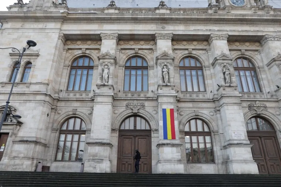 O persoană stă în fața unei uși de lemn de la o clădire monumentală cu ferestre arcuite și statui în nișe.
