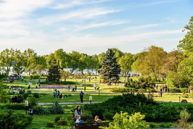 Oamenii se plimbă și stau pe iarbă într-un parc verde cu copaci și tufișuri sub un cer înzăpezit de nori albi.