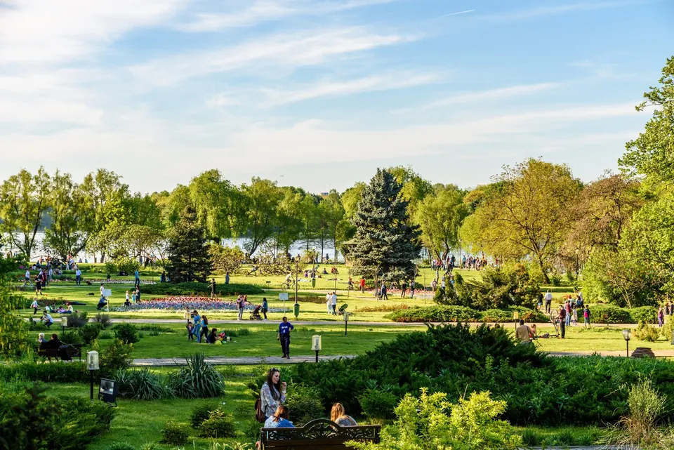 Oamenii se plimbă și stau pe iarbă într-un parc verde cu copaci și tufișuri sub un cer înzăpezit de nori albi.