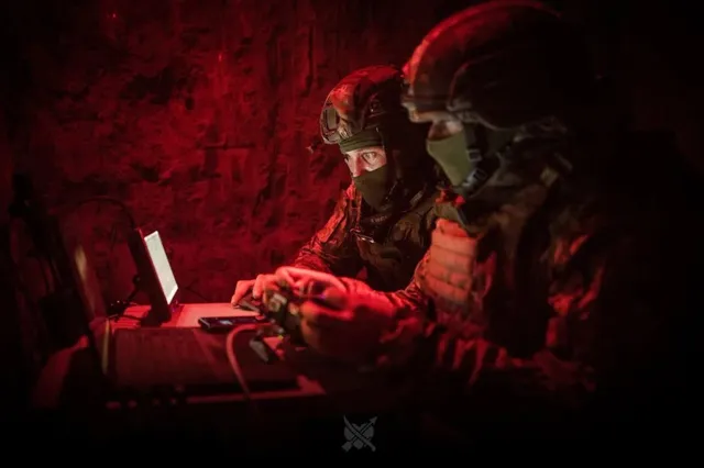 Două persoane purtând echipament militar și căști operează un laptop într-un mediu întunecat iluminat de o lumină roșie.