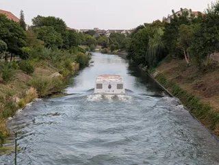 Un vaporaș alb navighează pe un canal înconjurat de vegetație și maluri înclinat, lăsând în urmă un urme de apă agitată.