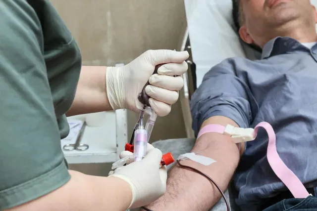 O persoană cu mâneca ridicată stă întinsă în timp ce un medic cu mănuși albe manipulează tuburi de recoltare a sângelui.