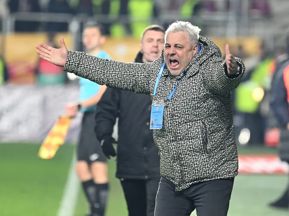 Un bărbat cu părul alb și o jachetă cu model geometric stă cu brațele întinse și gura deschisă pe un teren de fotbal.