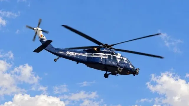 Un elicopter albastru cu marcaje albe și inscripția POLITIA zboară pe un cer albastru cu nori albi.