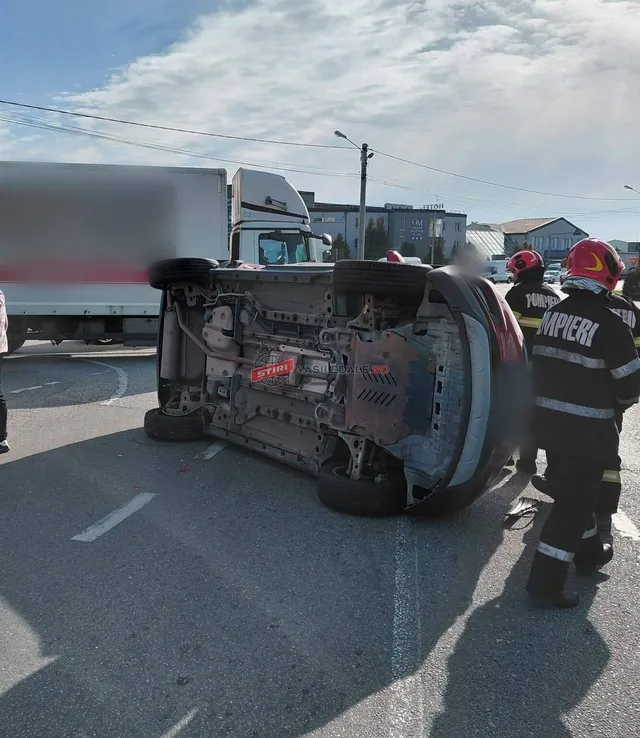 O mașină neagră este răsturnată pe o parte pe asfaltul unei străzi, lângă un camion și doi pompieri cu echipament de protecție.