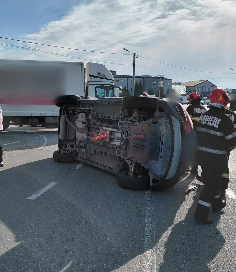 O mașină neagră este răsturnată pe o parte pe asfaltul unei străzi, lângă un camion și doi pompieri cu echipament de protecție.
