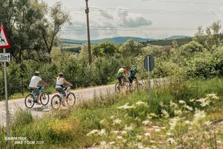 Patru persoane pe biciclete circulă pe un drum asfaltat înconjurat de vegetație verde și dealuri sub un cer înnorat.
