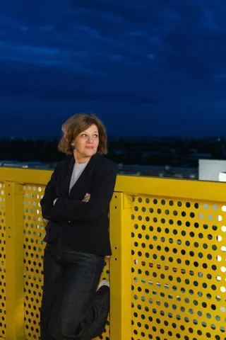 O femeie cu părul scurt și casetă neagră stă cu brațele încrucișate lângă un gard galben perforat sub un cer albastru închis.