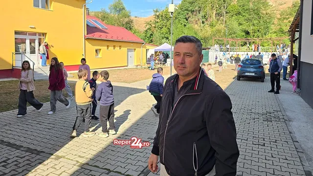 Un bărbat într-o jachetă neagră stă în prim-plan, în timp ce copii și alți adulți se află pe o curte pavată lângă clădiri colorate.
