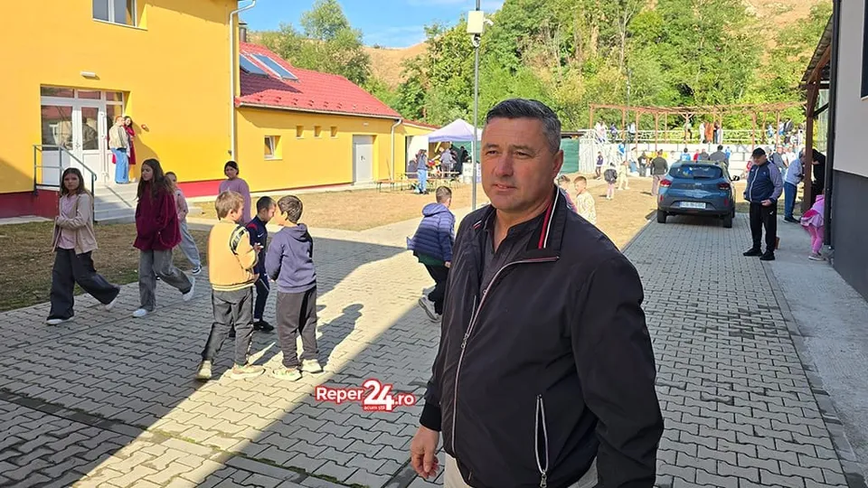 Un bărbat într-o jachetă neagră stă în prim-plan, în timp ce copii și alți adulți se află pe o curte pavată lângă clădiri colorate.
