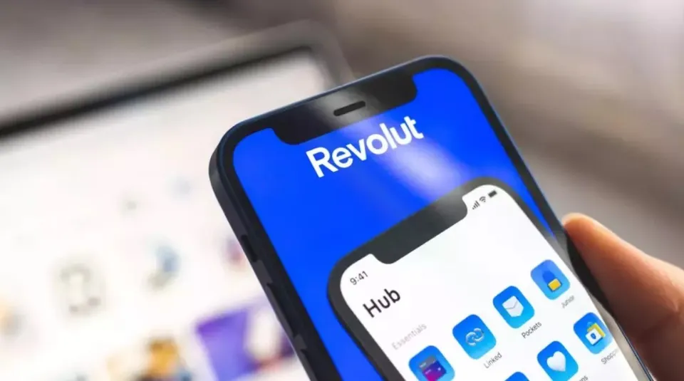 O mână ține un smartphone care afișează logo-ul Revolut pe un ecran albastru și o interfață de aplicație cu diverse pictograme colorate.