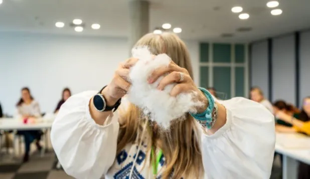 O persoană cu părul blond ține o mână de vată albă și pufoasă în fața feței, purtând o bluză albă cu mâneci largi.