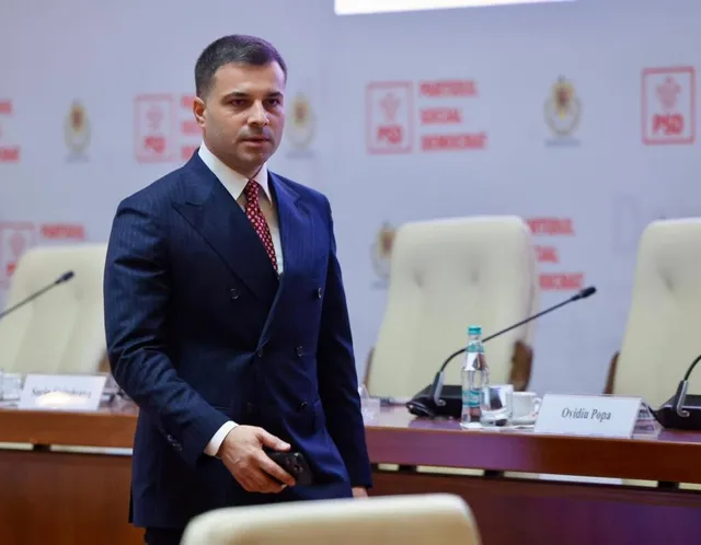 Un bărbat într-un costum albastru cu două rânduri de nasturi stă în picioare lângă un birou cu scaune albe și microfoane.