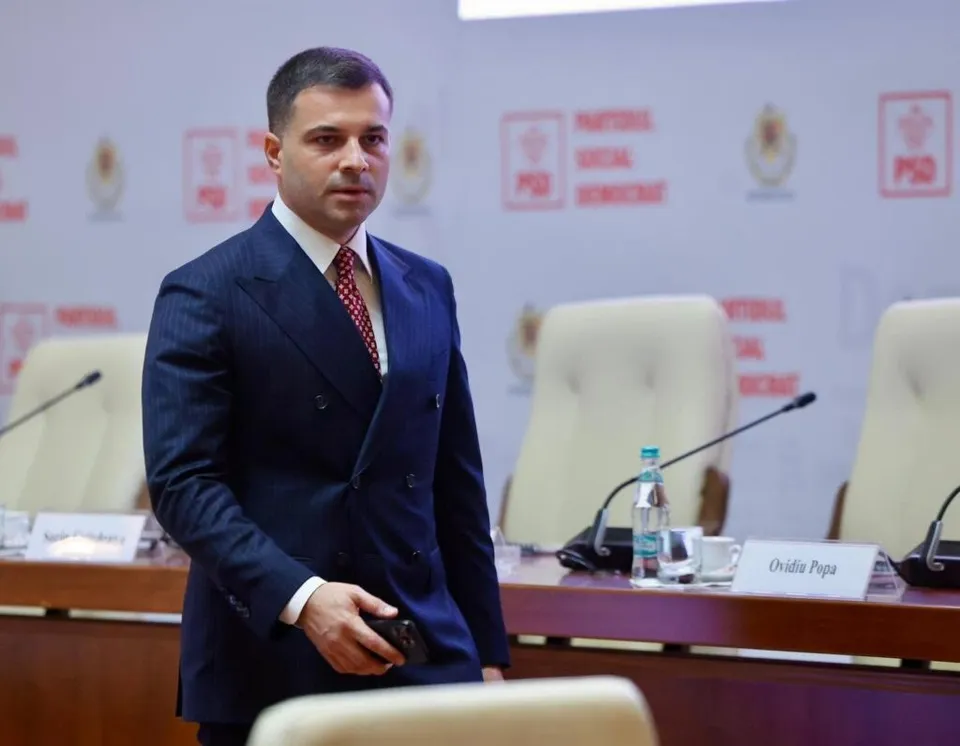 Un bărbat într-un costum albastru cu două rânduri de nasturi stă în picioare lângă un birou cu scaune albe și microfoane.