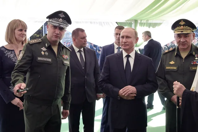 Un grup de bărbați în costume și uniforme militare stau în picioare sub o cortină de culoare deschisă.