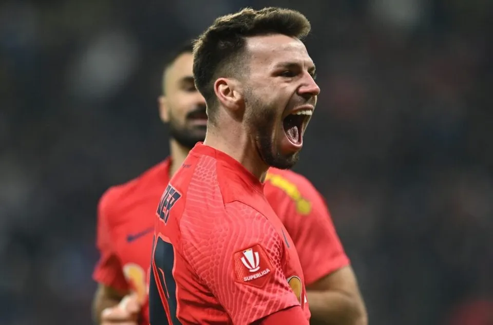 Un bărbat cu părul scurt și barbă poartă un tricou roșu și țipă cu gura larg deschisă într-un stadion.