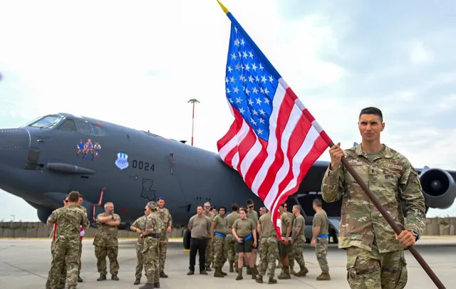 Un soldat în uniformă de camuflaj ține un steag american în fața unui avion militar gri și a unui grup de militari.