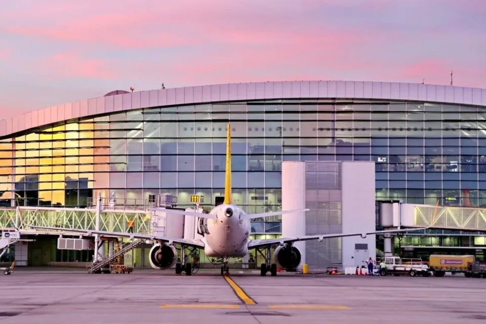 Un avion de pasageri este poziționat pe pista unui aeroport în fața unei clădiri terminale cu fațadă de sticlă sub un cer roz.