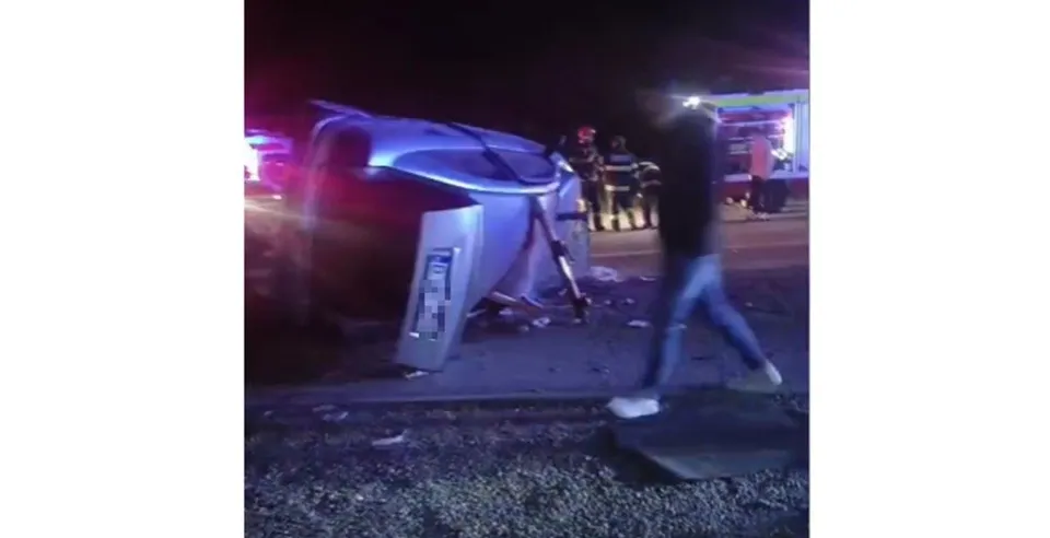 Un autoturism gri este răsturnat pe o parte pe marginea drumului, lângă un om care merge pe lângă vehicul noaptea.