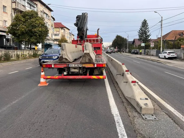 Un utilitar roșu transportă bariere de beton pe un drum asfaltat lângă o linie albă și un parapet de beton.